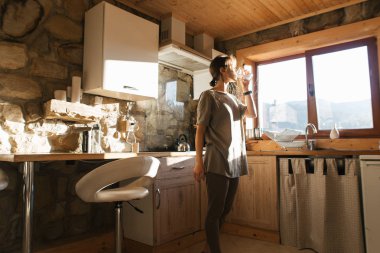 Genç bir kadın dağlardaki rahat mutfakta su içiyor.