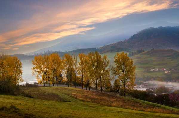 Mountain road with trees along a path with a mountain village in sunrise autumn morning. — Stock Photo, Image