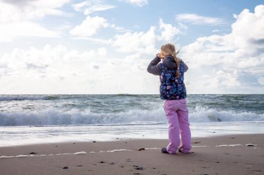Kumlu sahilde oynarken sevimli küçük kız. Sıcak çiçek baskı ceket, pom pom şapka ve açık havada Güz, kış veya bahar gününde oynarken eşarp giyen mutlu çocuk. Doğada kış yürüyüş zevk aile.