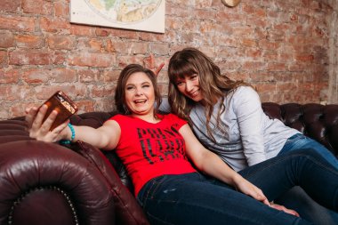 İki genç güzel neşeli kız mavi Jeans evde koltukta oturan gülen ve selfie yapmak