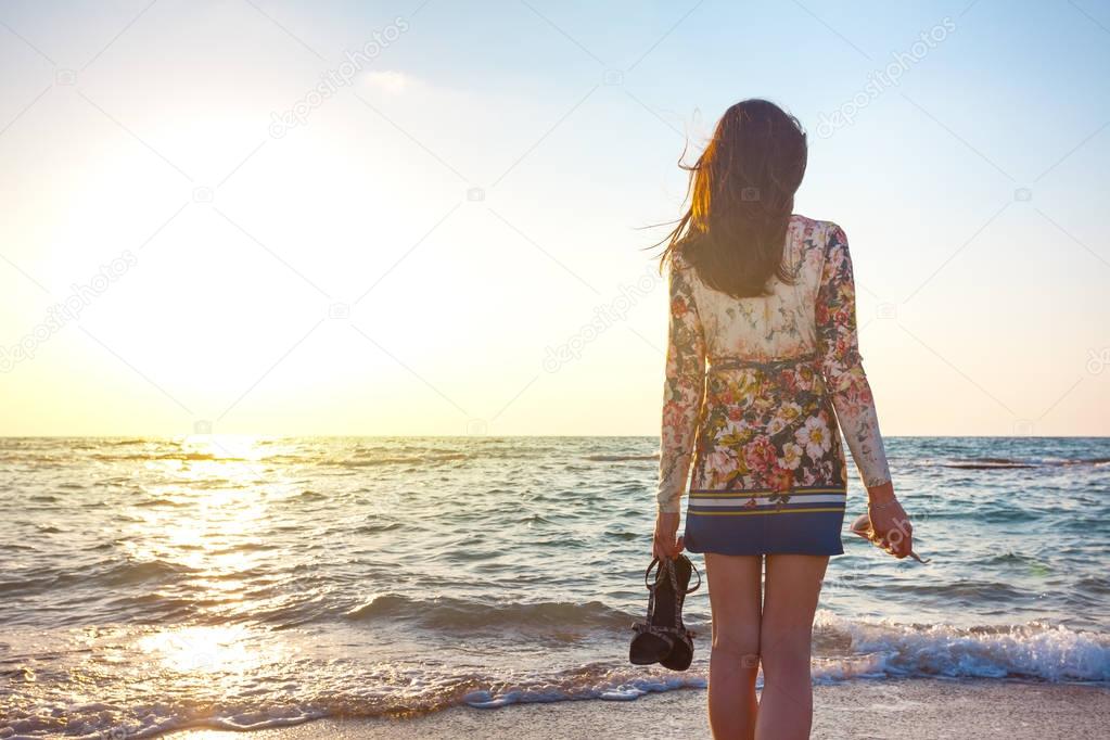 beautiful woman in colorful dress standing on the beach near the ocean and looking far away at the sunset