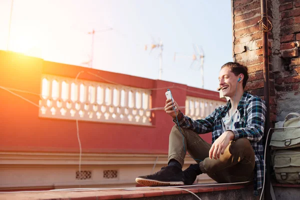 Усміхнений чоловік сидить на даху з мобільним телефоном та слухає музику з навушниками — стокове фото