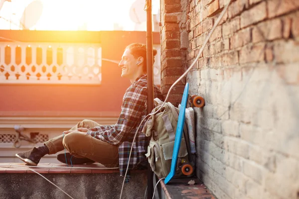 çatı ve sırt çantası ve kaykay ile kulaklık müzik dinleme kenarında oturan Gezgin adam gülümseyen