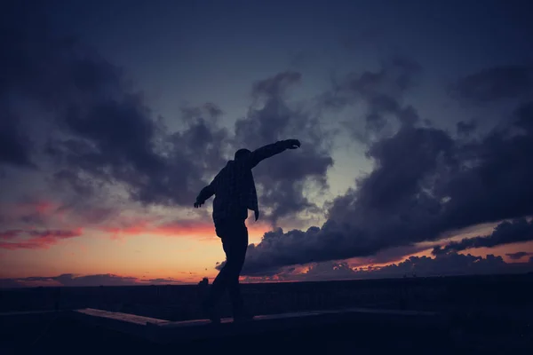 Silhueta de homem casual andando equilíbrio no topo do telhado na cidade ao pôr do sol — Fotografia de Stock