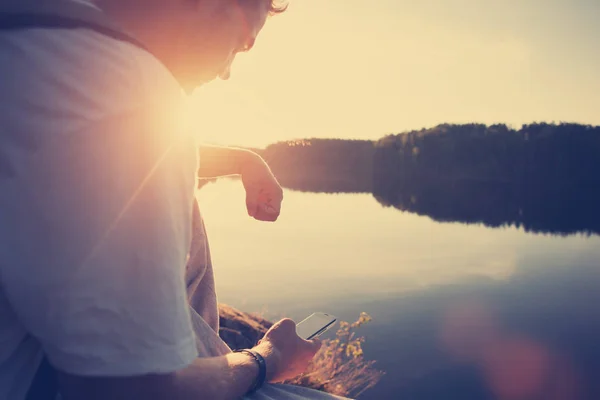 Voyageur homme assis au-dessus du lac au coucher du soleil avec téléphone portable dans ses mains — Photo