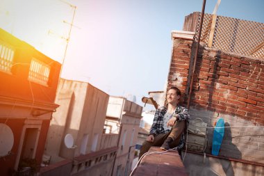 yüksek çatı uzakta görünümlü, sırt çantası ve kaykay ile kenarında oturan cesur adam