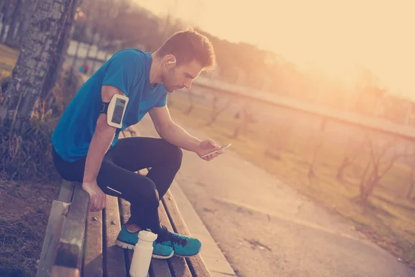 Hombre atleta sentado en el parque al atardecer con teléfono móvil, ar —  Fotos de Stock