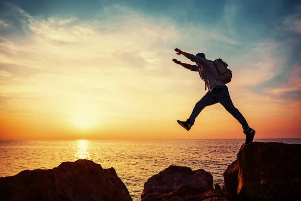 Giovane uomo coraggioso con zaino saltare sopra le rocce vicino al mare al tramonto — Foto Stock