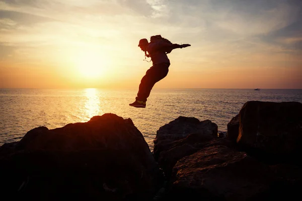 Brave man with backpack jumping over rocks near ocean in beautiful sunset — Stock Photo, Image