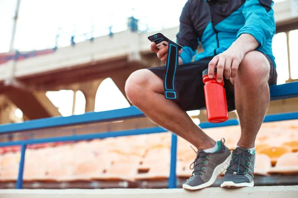 Αθλητής που κάθεται με μπουκάλι νερό και κινητό τηλέφωνο στο κιγκλίδωμα για το γήπεδο — Φωτογραφία Αρχείου