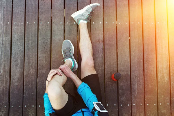 Jambes de sportif reposant assis sur le sol en bois avec bouteille en plastique sport rouge, vue du haut — Photo