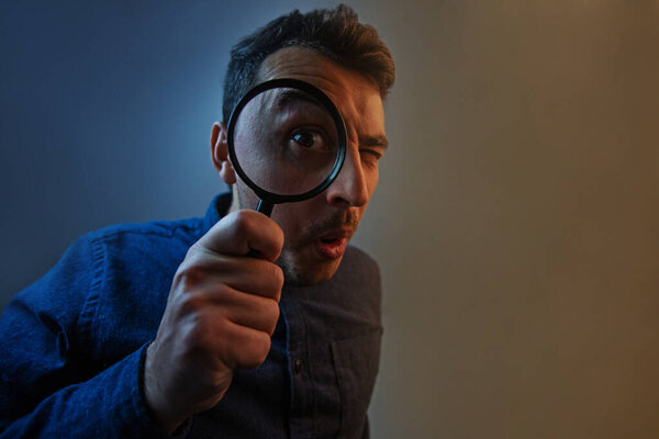 Shocked man with magnifying glass on grey background. Curious yo