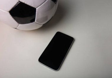 Canlı futbol bahis konsepti, boş akıllı telefon ekranı ve gri masa minimalist arka planında izole edilmiş klasik futbol topu. Spor kumar uygulaması, cep telefonuyla eğlenceli oyunlar, kopyalama alanı..