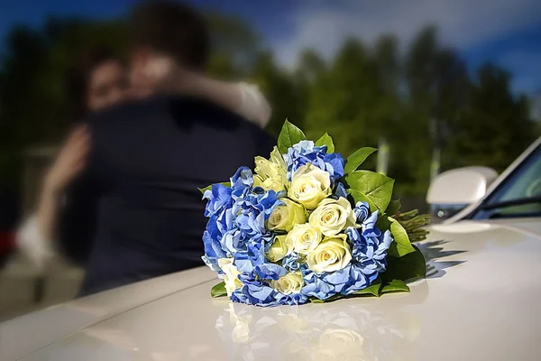 Bridal bouquet on a wedding car. In the background hugging the bride and groom blurry — Stock Photo, Image