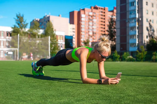 Sport e stile di vita. Slim fitness giovane ragazza con coda di cavallo facendo esercizio di fasciatura al chiuso sullo stadio verde e giocare in smart phone allo stesso tempo — Foto Stock