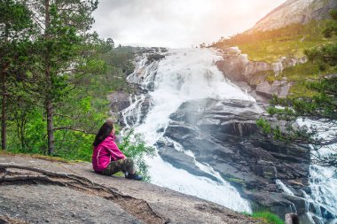 Taş üzerine oturan bir genç uzun yürüyüşe çıkan kimse kız ve şelaleler Norveç vadisinde bir şelale hayran