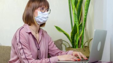 Evde kal ve gezegeni kurtar. Kadın yüzüne tıbbi bir maske takar ve dizüstü bilgisayarla çalışır. 4K 'da Coronavirus kontrolü.