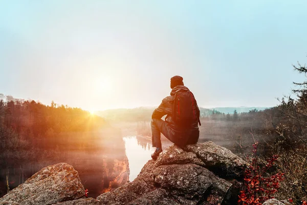 身穿夹克 裤子和背包的年轻人坐在岩石上 望着日落中的沃尔塔瓦河和山谷 — 图库照片