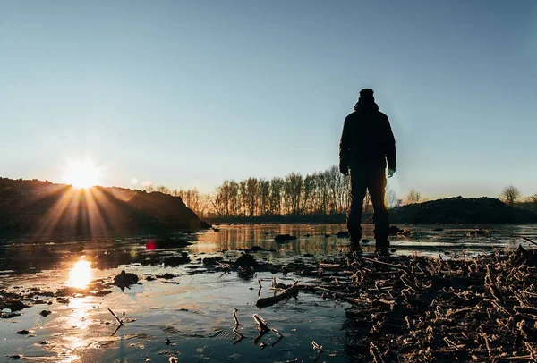 Молодий чоловік стоїть на льоду в ставку з замерзлим берегом, сонцем і деревом — стокове фото