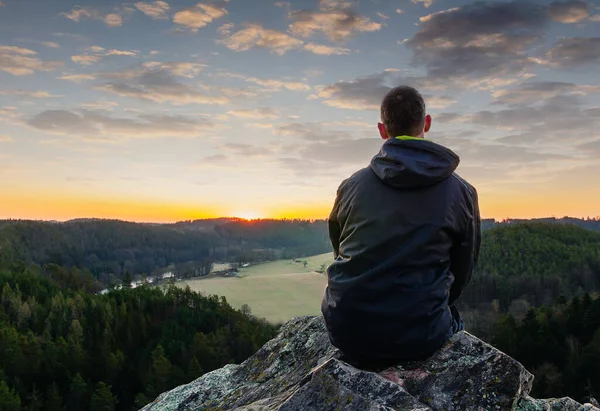 穿着夹克 坐在岩石上 望着日出时分的山谷的年轻人 — 图库照片