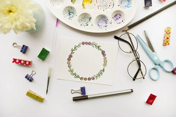 Artist\'s workspace. Floral wreath frame painted with watercolor, chrysanthemum, glasses, paintbrush, scissors, watercolor, palette on a white background. Flat lay
