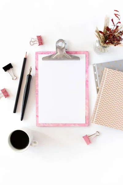 Home office desk with pink clipboard, notepads, bouquet of dry flowers, calligraphic pen, pencil, paper clips, mug of coffee on a white background