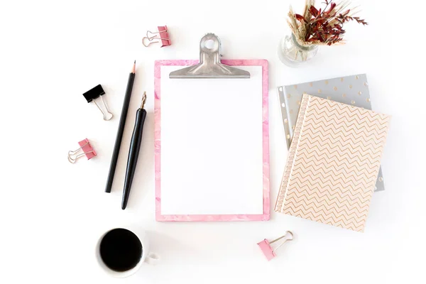 Home office desk with pink clipboard, notepads, bouquet of dry flowers, calligraphic pen, pencil, paper clips, mug of coffee on a white background