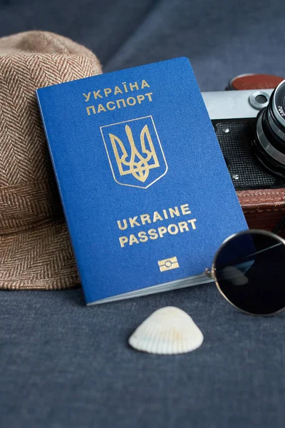Pasaporte de viaje ucraniano sobre fondo gris. gafas de sol, sombrero. cámara vintage en el fondo. Acceso sin visado a la UE. Profundidad superficial del campo, enfoque en el logotipo ucraniano en el pasaporte . — Foto de Stock