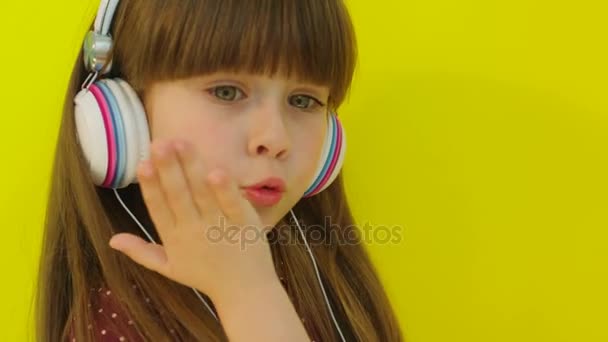 Gadis kecil yang cantik memakai headphone putih dan ciuman di depan kamera. Latar belakang kuning. Tutup. — Stok Video