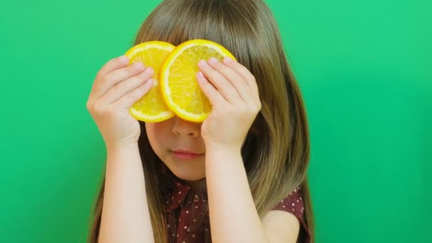 Linda niña sosteniendo una rebanada de naranja cerca de sus ojos y escondiéndose. Fondo verde — Vídeos de Stock