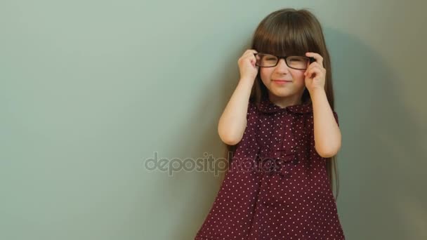 Мила дівчинка позує і посміхається в камеру. Маленька дівчинка в окулярах. Портрет знятий. Сірий фон — стокове відео