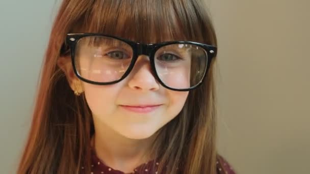 Забавная маленькая девочка надевает большие взрослые очки. Маленькая девочка развлекается на камеру. Закрывай. Серый фон — стоковое видео