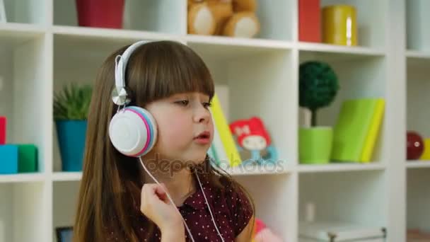 Cute little girl dancing and singing while listening to music in white headphones. Furniture background — Stock Video