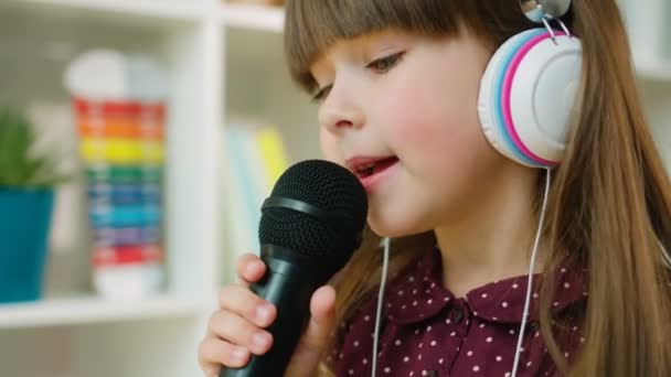 Крупним планом мила дівчинка співає на мікрофоні в білих навушниках. Маленька дівчинка переїжджає в туалет. Внутрішня стрільба — стокове відео