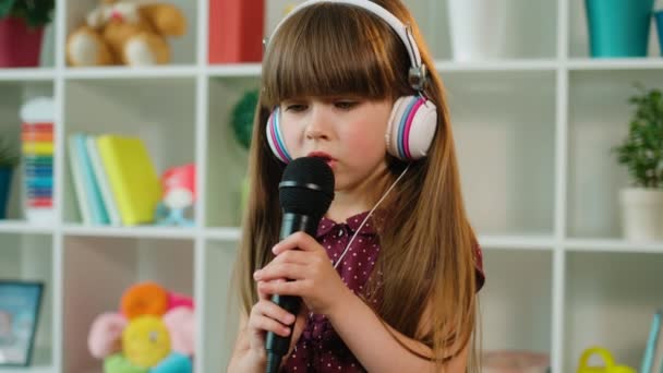 Κοντινό πλάνο όμορφο κοριτσάκι, τραγουδώντας για το μικρόφωνο φορώντας λευκά ακουστικά. Μικρό κορίτσι που κινείται προς το rytm του τραγουδιού. Εσωτερική γυρίσματα — Αρχείο Βίντεο