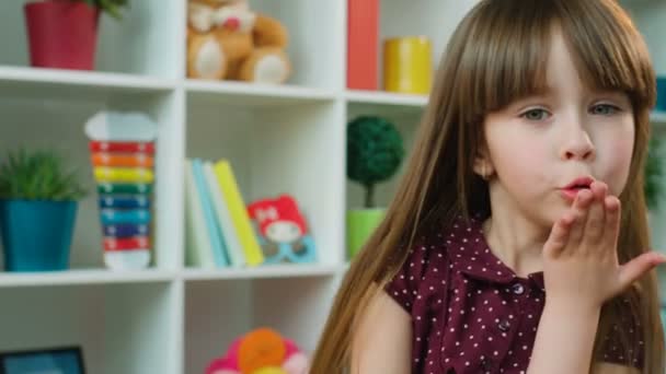 Cute little girl blowing kisses at the camera in kids room. White shelves with toys background. Close up — Stock Video