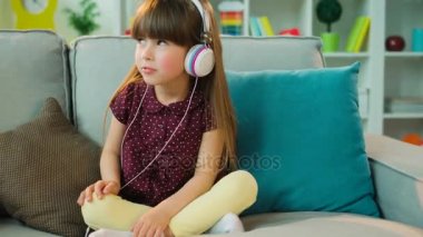 Büyük beyaz kulaklık müzik ve dans, lotus pozisyonda oturma odasında kanepede otururken sevimli küçük kız. Yakın çekim