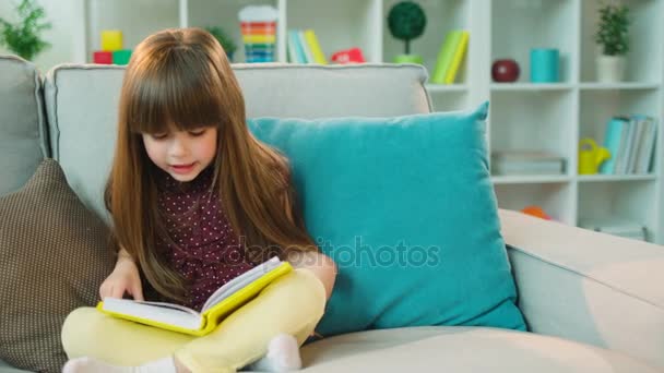 Mooi klein meisje het lezen van interessante boek zittend op de Bank in de lotuspositie in de woonkamer. — Stockvideo