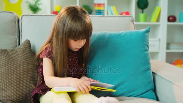 Gadis kecil yang tampan duduk di sofa di ruang tamu dan membaca buku. Tertarik. Tembakan dalam ruangan — Stok Video