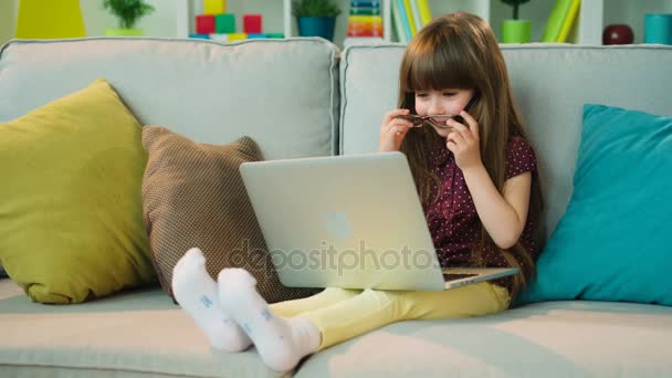 Linda niña usando un ordenador portátil en el sofá de la sala de estar. Grandes vasos cayendo de su cabeza. Gracioso. . — Vídeos de Stock