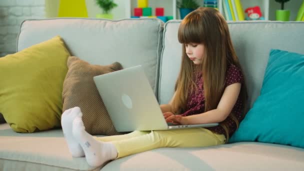 Söt liten flicka funderar på något och skriva ner det i bärbar dator. Lilla flickan har en idé. Sitter i soffan — Stockvideo