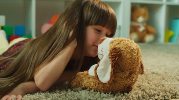 Pretty little girl lying on the floor and kissing and hugging her teddy bear. Close up — Stock Video
