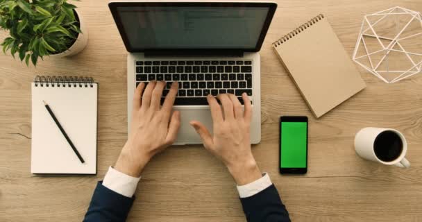 木製オフィス デスクでのノート パソコンで書いているビジネスその男。グリーン スクリーンのスマート フォン。手平面図です。スローモーション。赤の叙事詩 — ストック動画