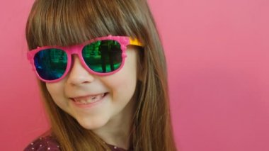 Yüz portre seyir ve kameraya gülümseyen pembe gözlüklü şirin küçük kız. Pembe bir arka plan. Yakın çekim