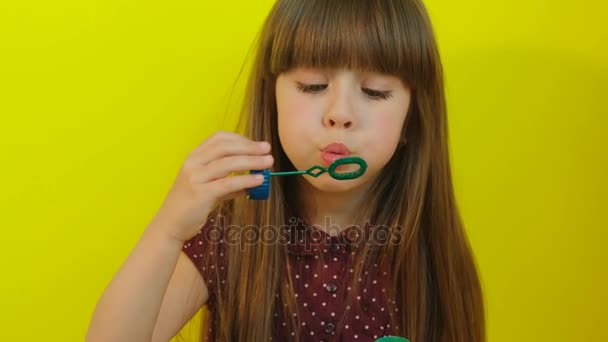 Hermosa niña soplando burbujas de jabón en la cámara. Fondo amarillo. Chica divirtiéndose. De cerca. — Vídeos de Stock