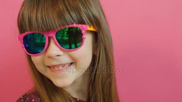 Красивый портрет симпатичной маленькой девочки в розовых очках, смотрящей и улыбающейся в камеру. Розовый фон. Закрыть — стоковое видео