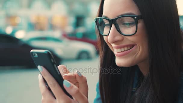 Mujer atractiva joven en gafas usando teléfono inteligente, tocando en la pantalla táctil, sonriendo mientras está sentado en las escaleras afuera en la calle de la ciudad. De cerca. — Vídeos de Stock