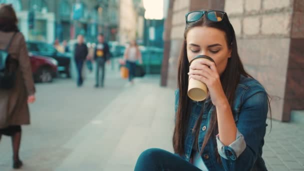 Красивая молодая женщина сидит на ступеньках на городской улице и пьет кофе. Женщина смотрит в камеру и улыбается — стоковое видео