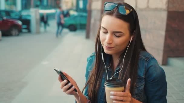 O tânără frumoasă ascultă muzică pe telefonul inteligent, cântă și se mută în ritm în timp ce stă pe treptele de afară de pe stradă. Close up — Videoclip de stoc