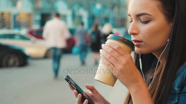 Красивая молодая женщина слушает йогурт по смартфону, переходит в ритм и пьет кофе, сидя на ступеньках на улице. Закрыть — стоковое видео